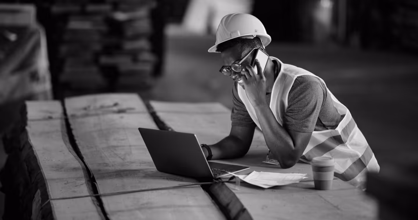 Man wearing a safety helmet and vest talking on a phone while looking at a laptop on a wooden table in an industrial setting.