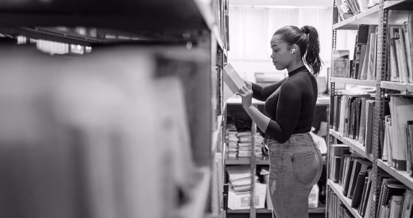 Woman with ponytail wearing earbuds and jeans selecting a book from a library shelf.
