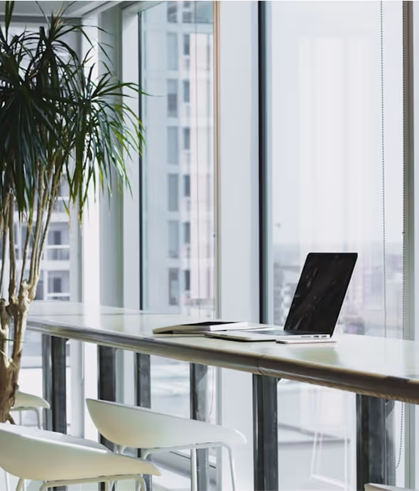 Modern workspace with a laptop and open book on a long table next to large windows and white chairs.
