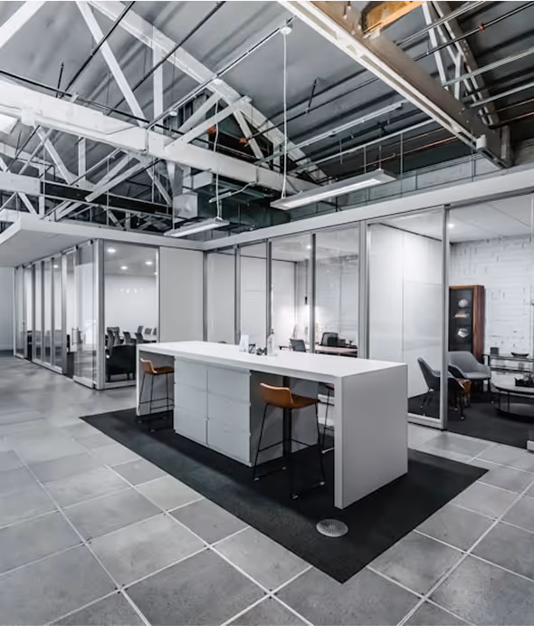 Modern office interior with glass-walled rooms, a white island desk with two brown stools, and exposed ceiling beams.