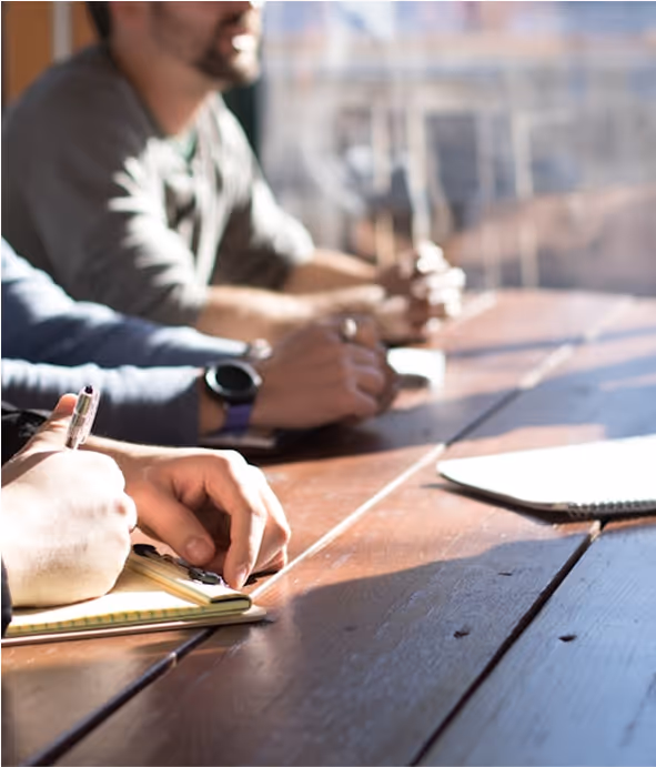 People sitting at a wooden table, writing notes on notepads with pens in a well-lit room.