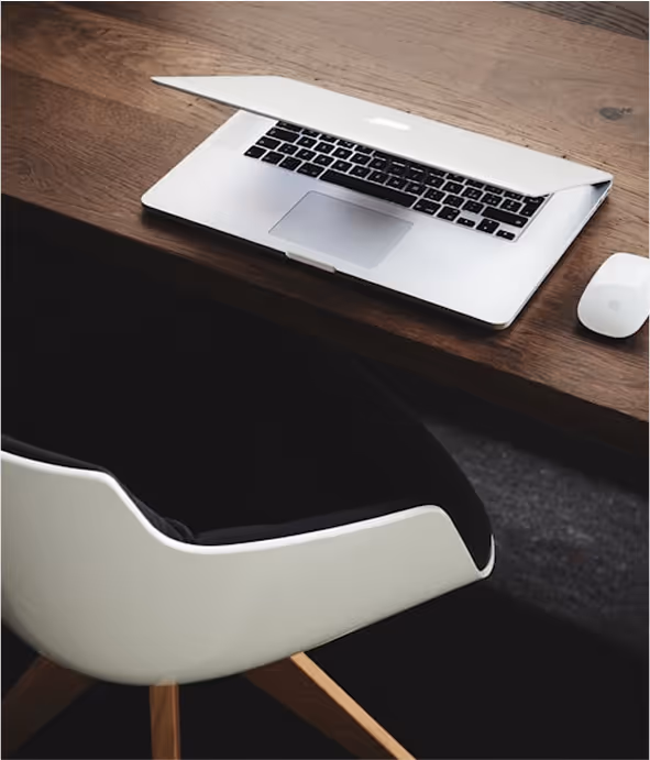 Partially closed silver laptop and white mouse on wooden desk next to white and black chair.