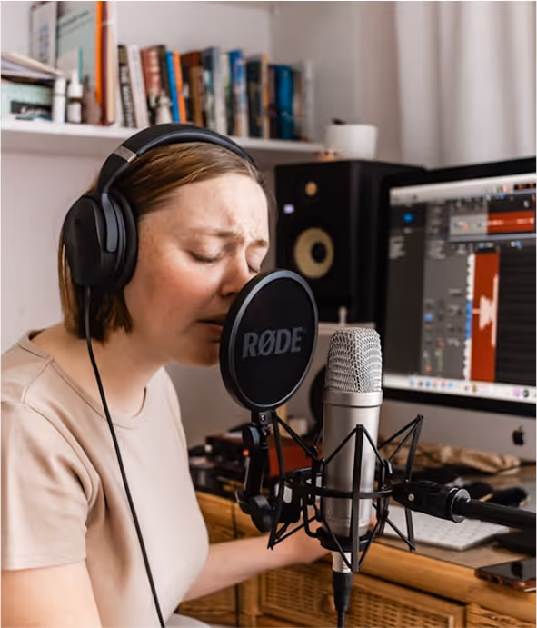 Person wearing headphones and singing into a RØDE microphone in a home recording setup with a computer displaying audio software in the background.