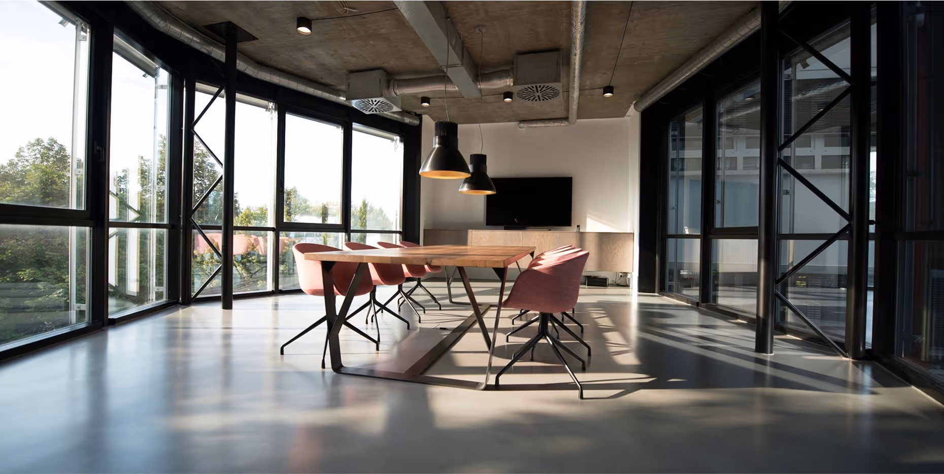 Modern office meeting room with large wooden table, pink chairs, two hanging lamps, and floor-to-ceiling windows.