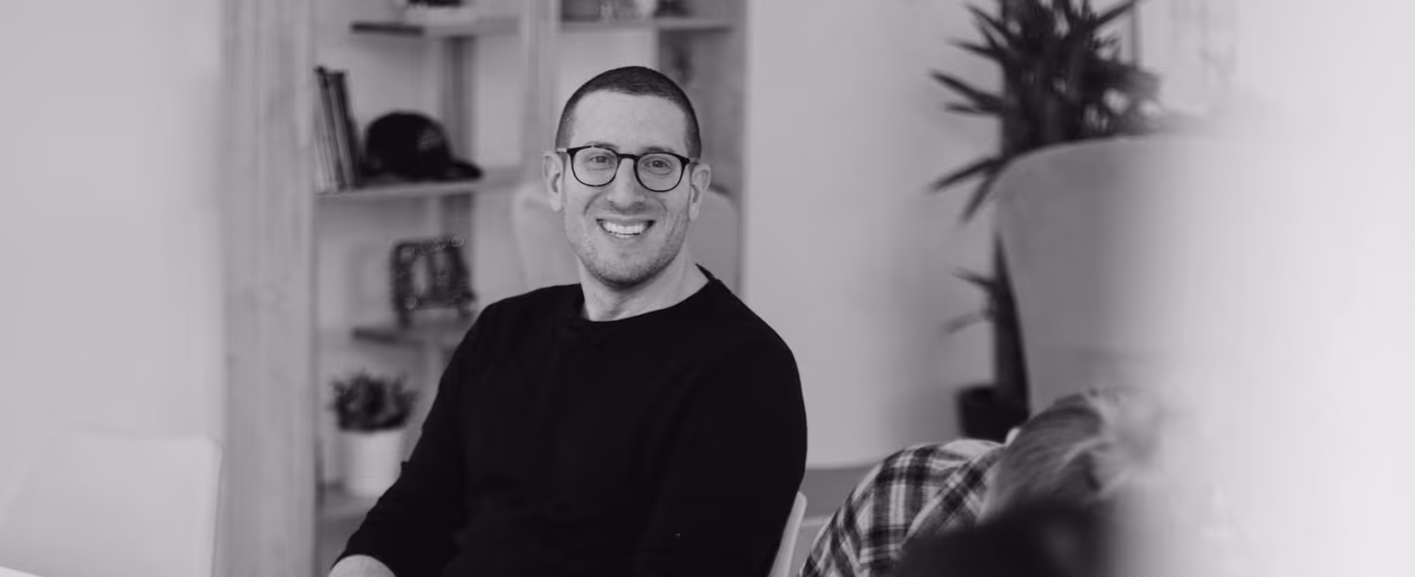 Smiling man wearing glasses and a dark sweater sitting in a room with shelves and plants.