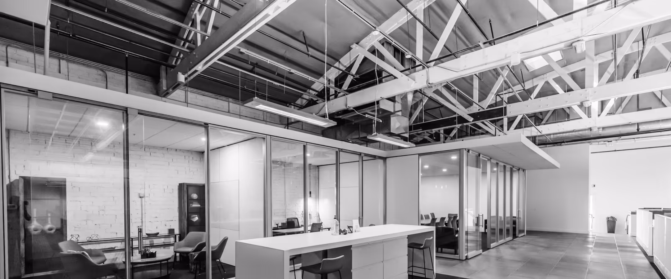 Modern office space with glass-walled meeting rooms, exposed ceiling beams, and a white central island with chairs.
