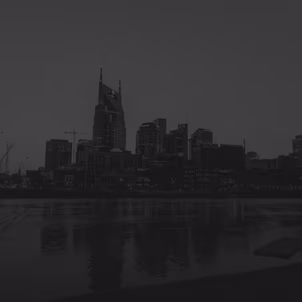 Black and white photo of a city skyline with tall buildings reflecting on a calm river at dusk.