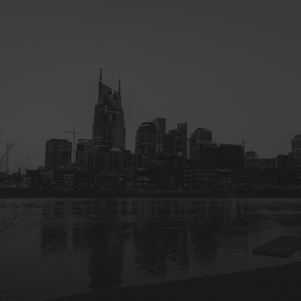 Black and white photo of a city skyline with tall buildings reflecting on a calm river at dusk.