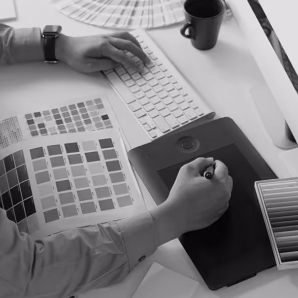 Person using a graphic tablet and keyboard with color swatch books and a coffee cup on a white desk.