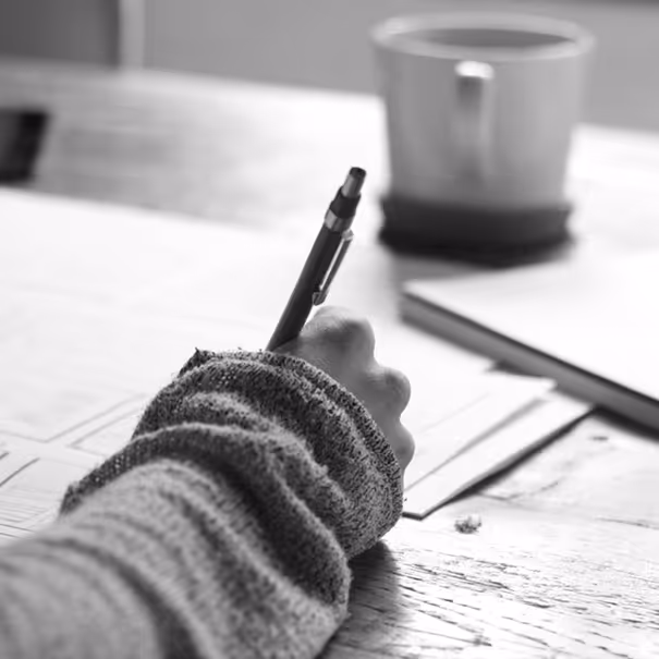 Hand holding a pen writing on paper at a wooden table with a blurred coffee cup in the background.