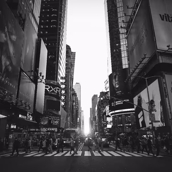 Black and white photo of a busy city street with people crossing at a zebra crossing and tall buildings with illuminated billboards on both sides.