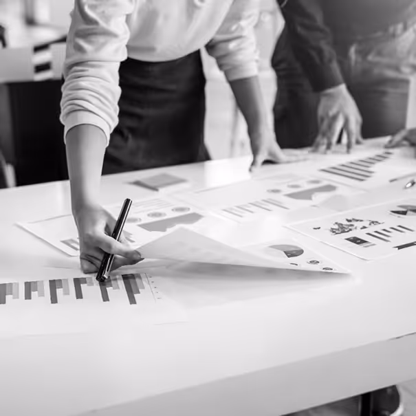 Two people reviewing business charts and graphs spread out on a white table, one person holding a pen and a sheet of paper.