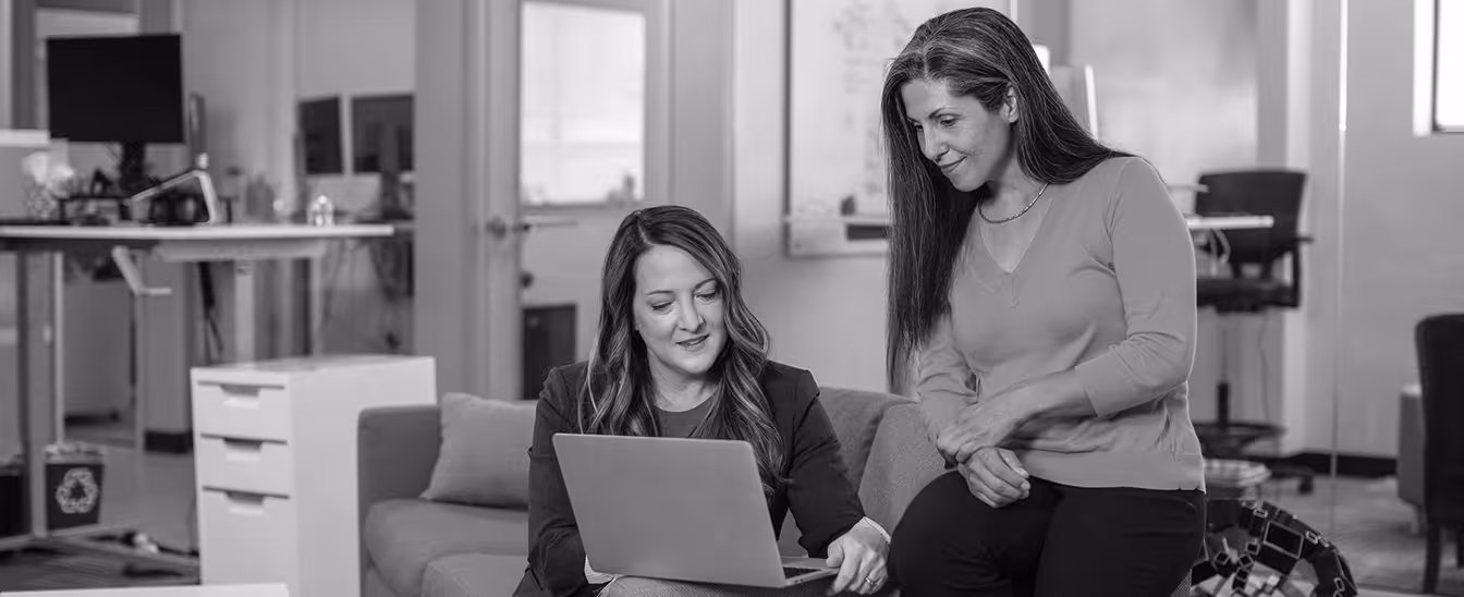 Two women collaborating in a modern office, one sitting on a couch showing a laptop screen to the other who is leaning on the couch.