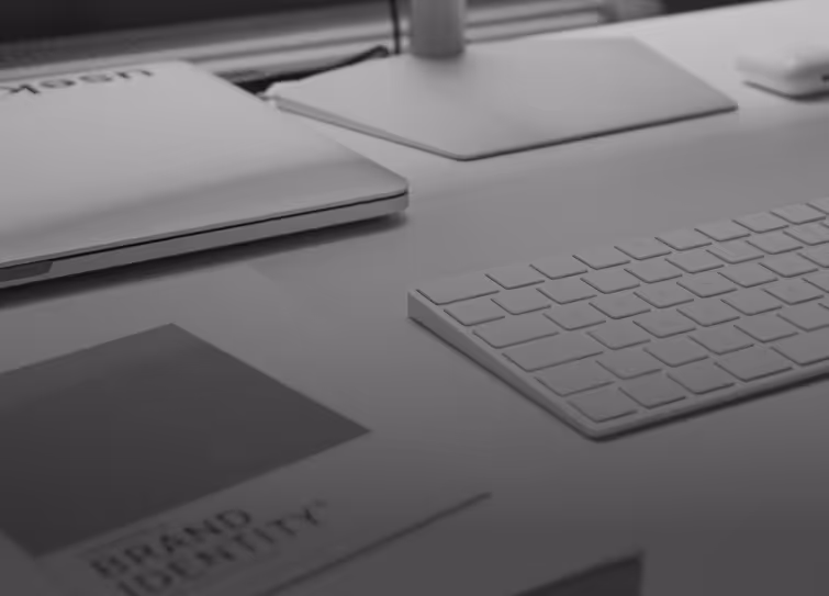 Close-up of a clean white desk setup with a wireless keyboard, laptop, monitor stand, and a booklet titled 'Brand Identity'.