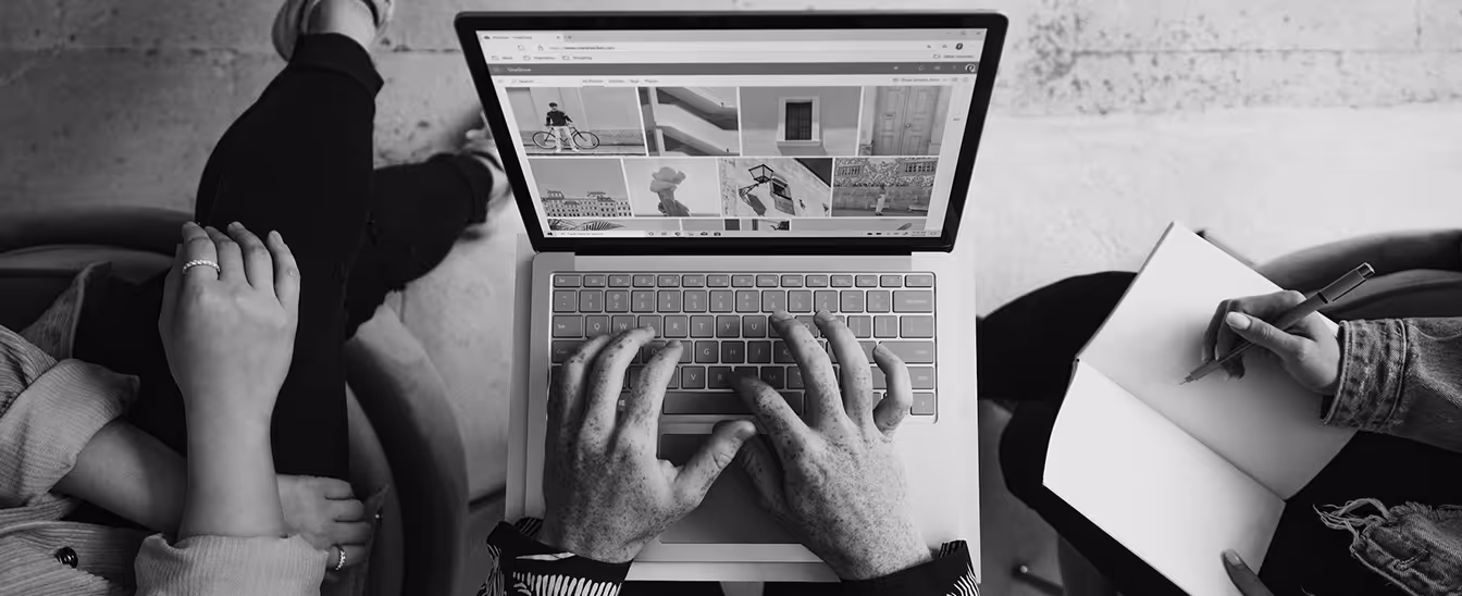 Overhead view of two people sitting on either side of a person typing on a laptop showing a webpage with photos, while one of them writes in a notebook.