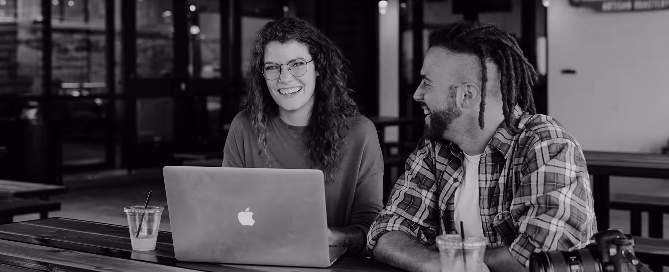 Two people sitting at a wooden table with a laptop and drinks, smiling and engaged in conversation indoors.