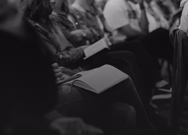 Black and white photo of people seated in rows, one person writing in an open notebook on their lap.