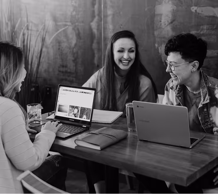 Three young adults sitting around a wooden table laughing and working on laptops in a casual indoor setting.