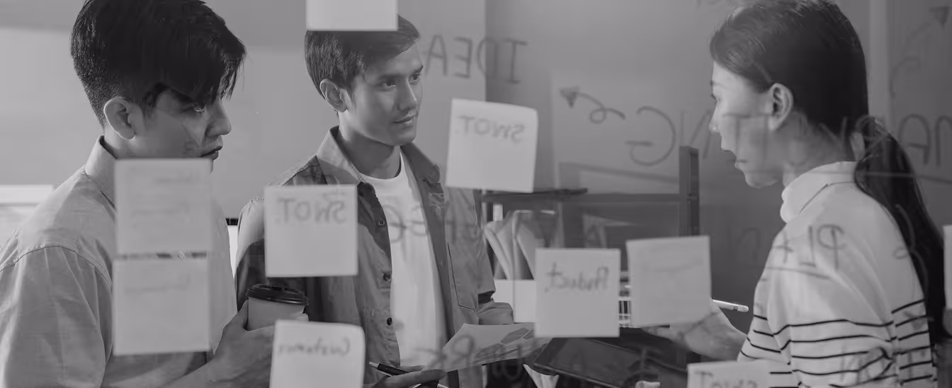 Three young professionals discussing and collaborating with sticky notes on a glass wall.
