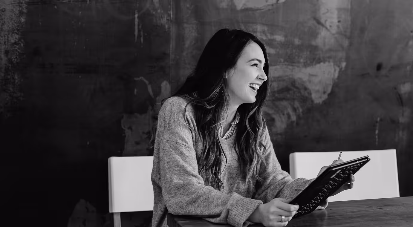 Smiling woman with long hair holding a tablet and stylus while sitting at a table against a dark textured wall.
