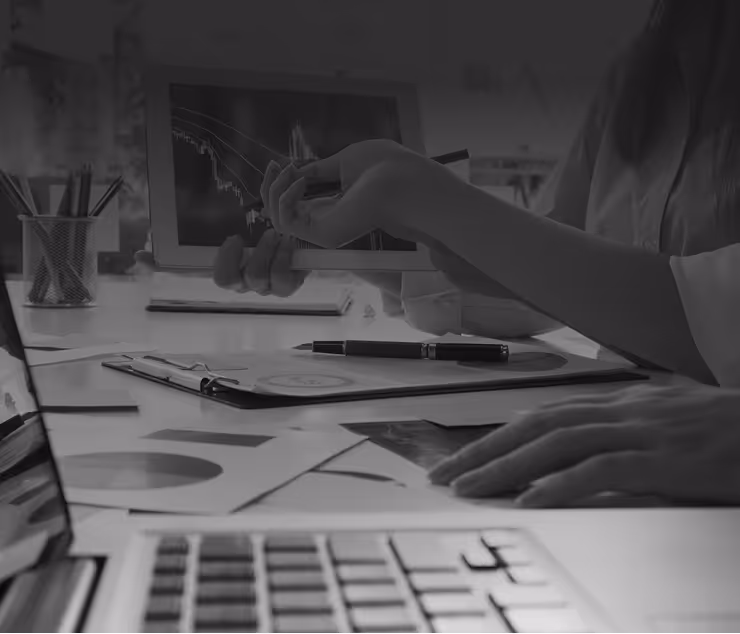Person pointing with a pen at a tablet displaying a downward trending financial chart while sitting at a desk with a laptop and documents.