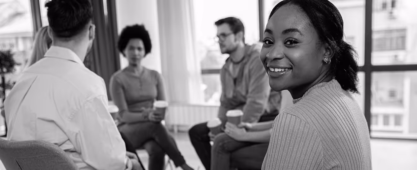 Group of diverse young adults sitting in a circle, holding coffee cups, with a smiling woman looking at the camera.