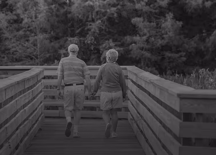 Elderly couple holding hands walking on a wooden boardwalk surrounded by trees.