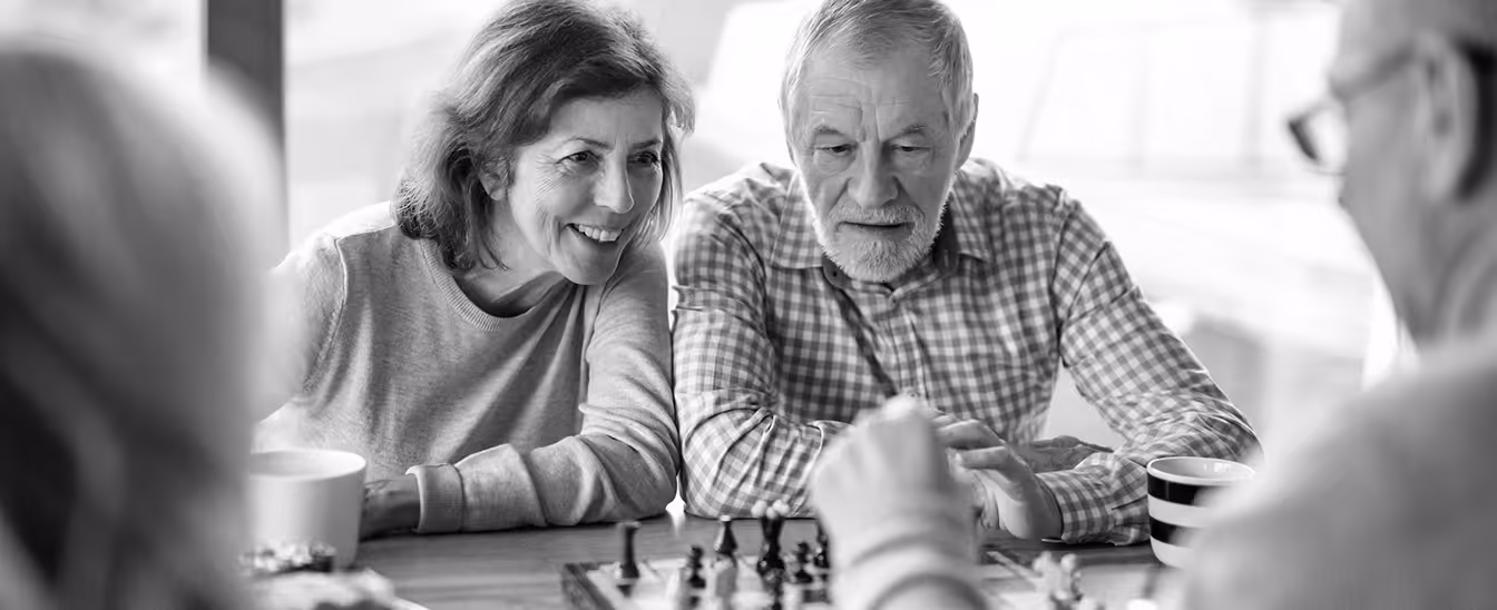 Two elderly adults, a woman and a man, smiling and watching a chess game at a table with coffee cups.