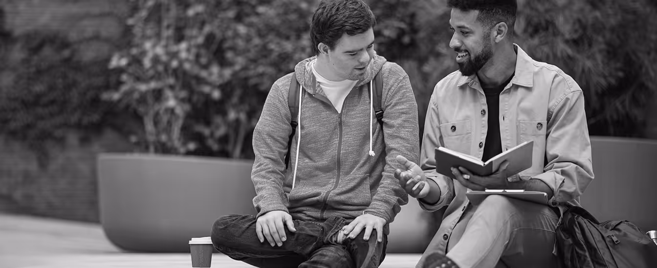 Two young men sitting outdoors, one holding an open book and the other listening attentively.