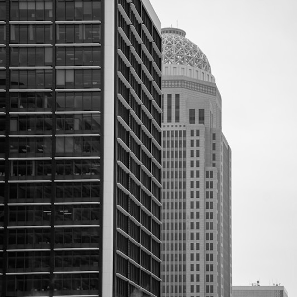 Black and white photo of two tall office buildings, one with a grid-like window pattern and the other with a rounded, patterned top.