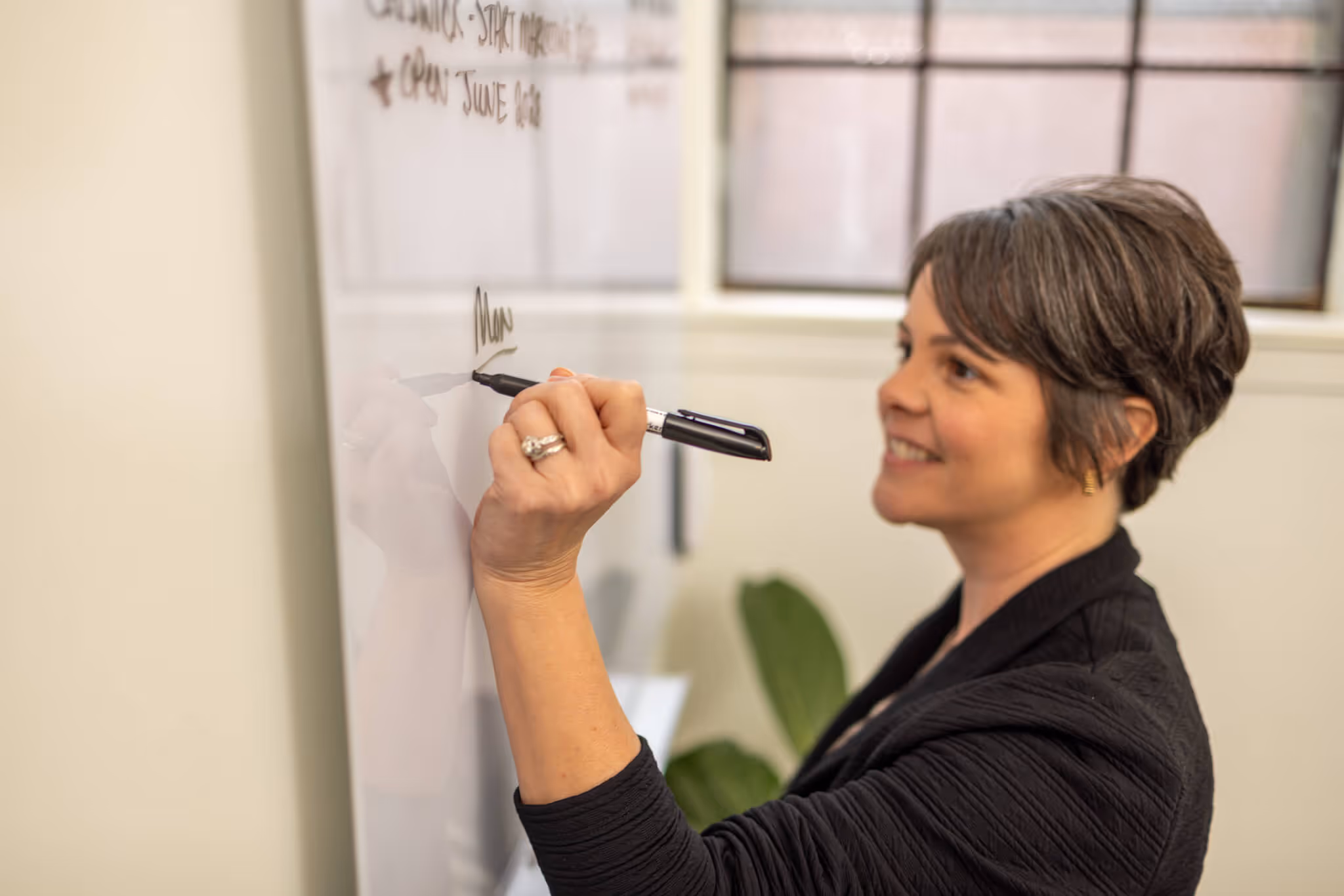 Two people sitting at a table covered with fabric samples and design papers, looking at a laptop together in a modern office with large windows.