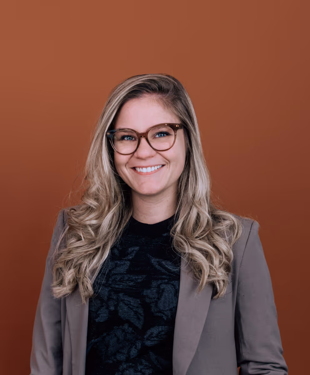 Femme souriante aux cheveux blonds bouclés, portant des lunettes et un blazer marron devant un fond uni orange.