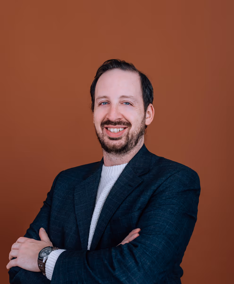 Homme souriant avec barbe et moustache, portant un blazer bleu foncé et un pull blanc, bras croisés devant un fond marron.