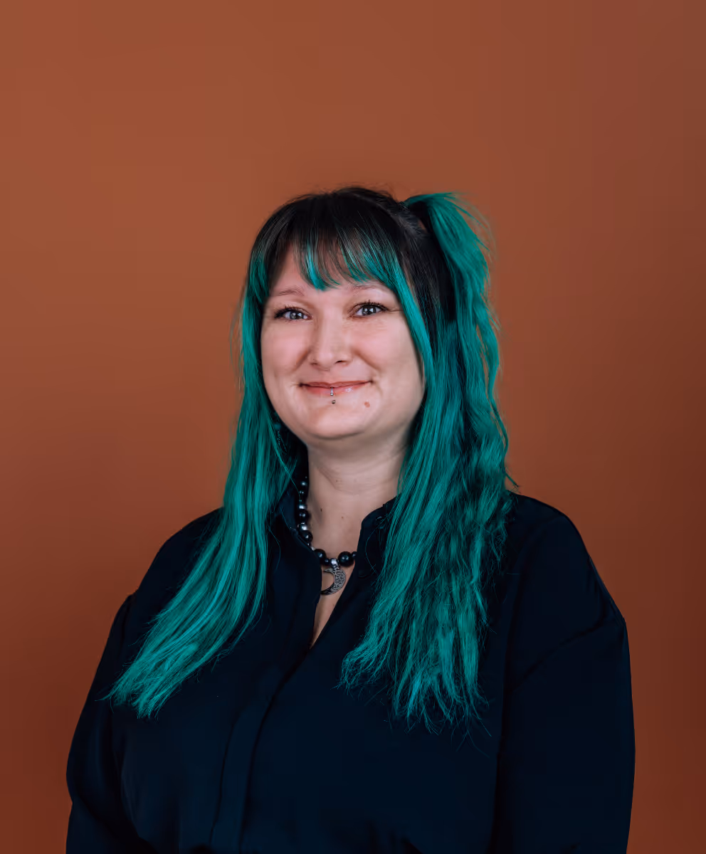 Portrait d’une femme avec des cheveux longs verts, portant une chemise noire et un collier avec un pendentif lune, sur un fond marron.