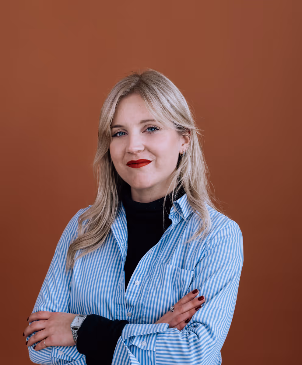 Femme blonde avec une chemise bleu et blanche à rayures, bras croisés, souriant devant un fond marron.