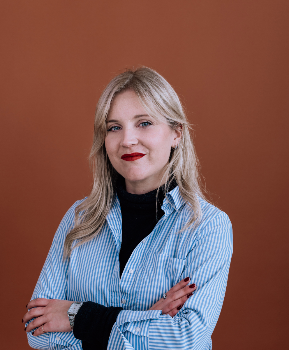 Femme blonde avec une chemise bleu et blanche à rayures, bras croisés, souriant devant un fond marron.