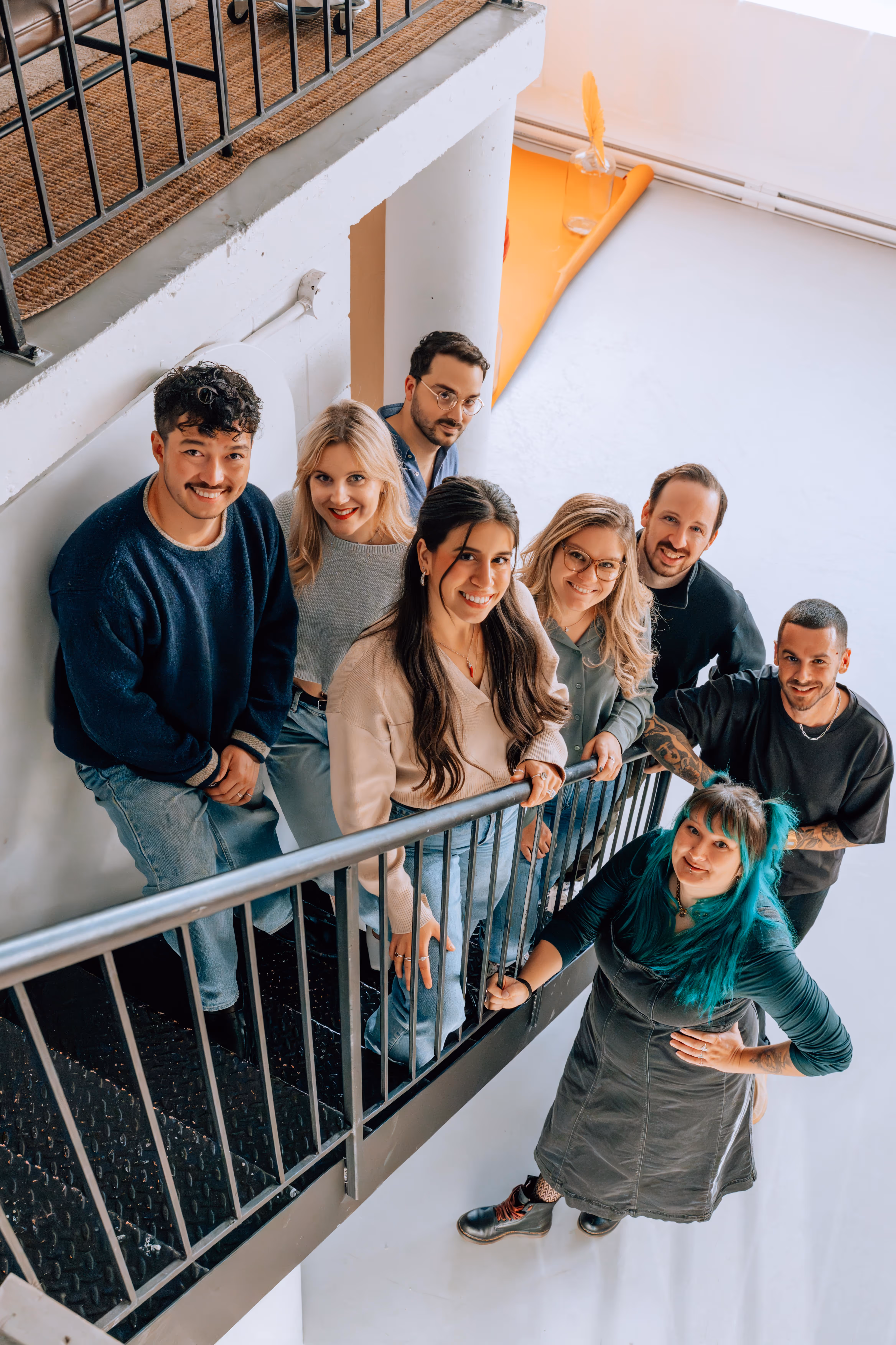 Groupe de huit jeunes adultes souriants sur un escalier intérieur en métal.