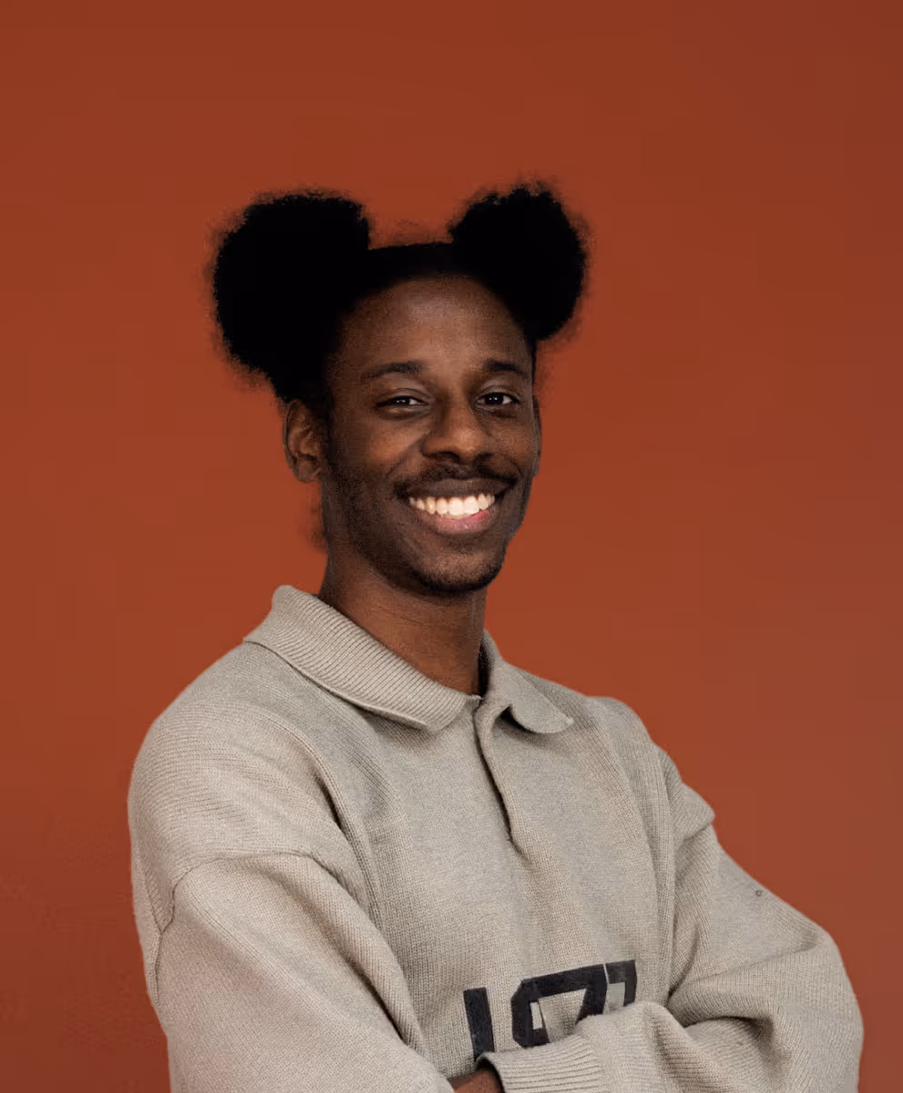 Homme souriant aux cheveux en deux boules, portant un pull gris, sur fond rouge.