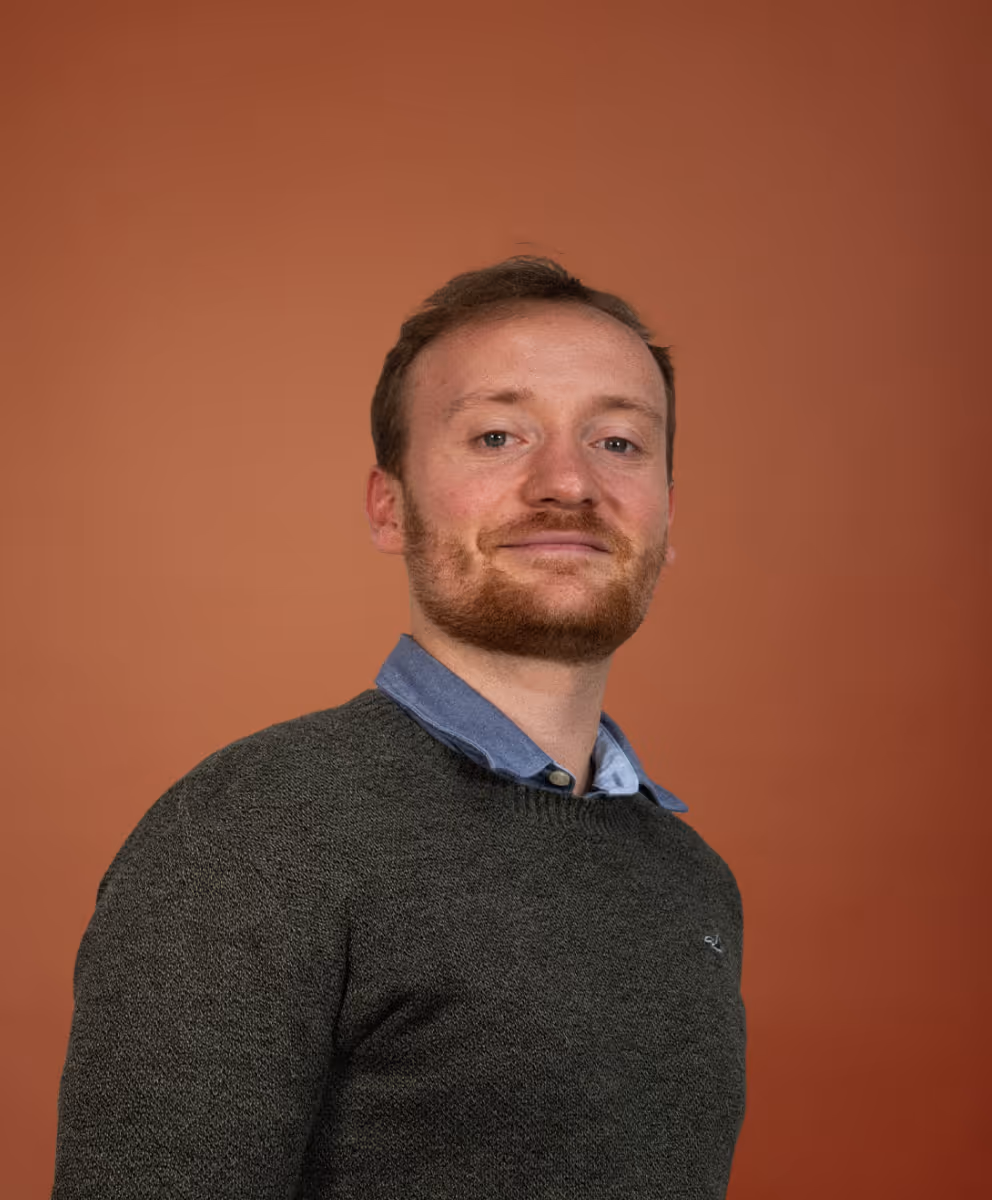 Homme avec cheveux roux et barbe portant un chandail gris et une chemise bleue, regardant vers l'avant avec un léger sourire devant un arrière-plan orange.