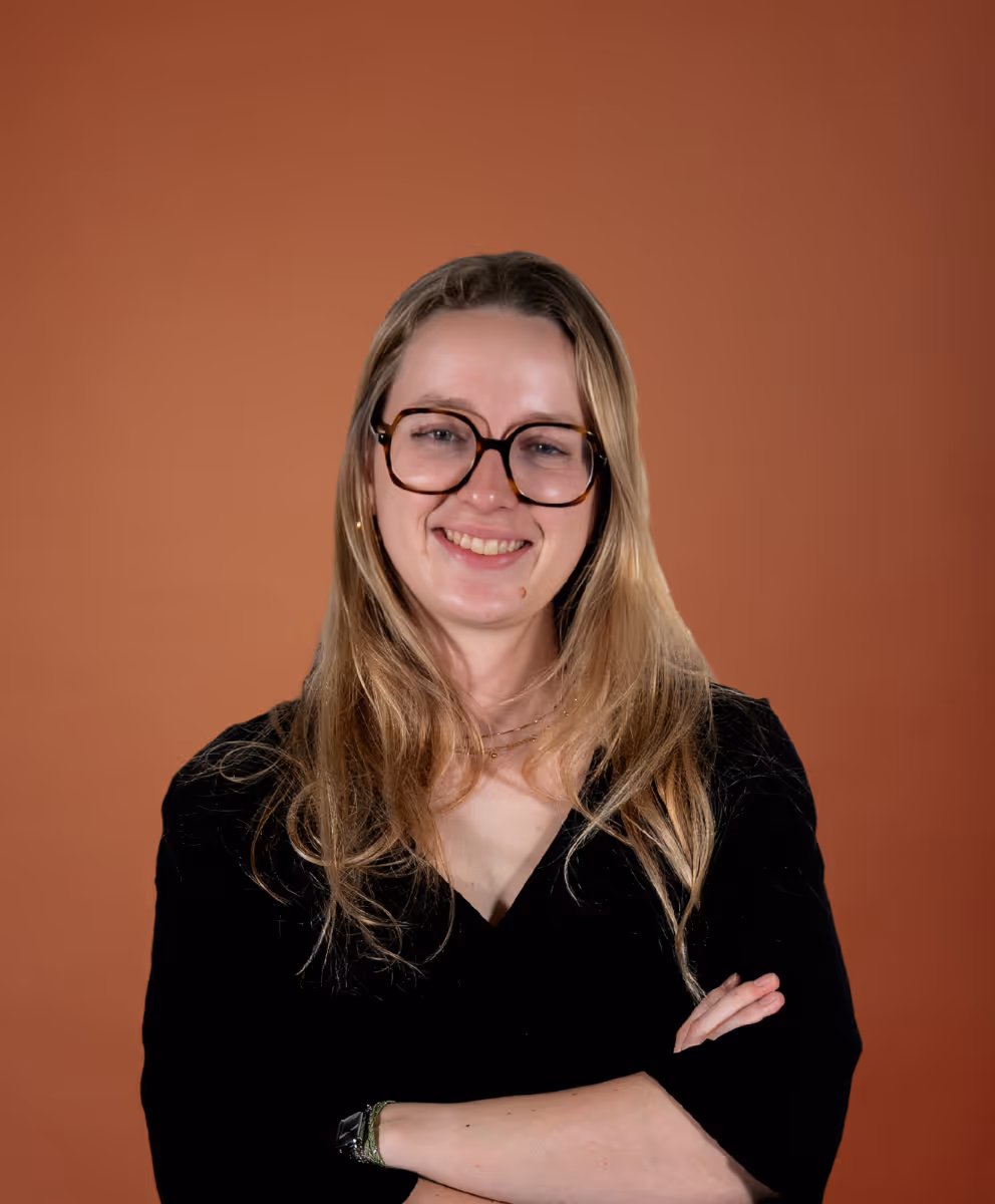 Jeune femme souriante avec lunettes et bras croisés devant un fond brun uni.