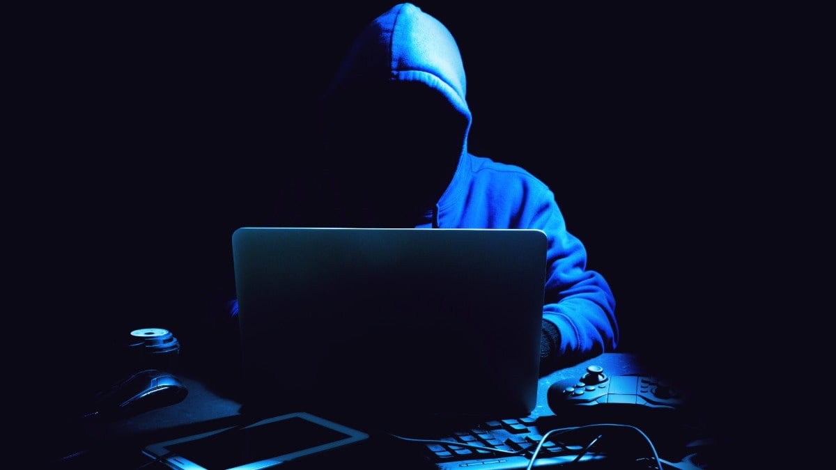 Dark blue concept image of a man in a hoodie with a computer committing identity theft through social engineering attacks