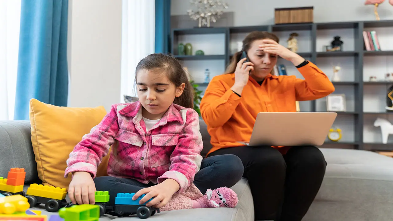 A concerned mother on the phone with a laptop open while her young daughter plays nearby, representing a parent dealing with child identity theft.