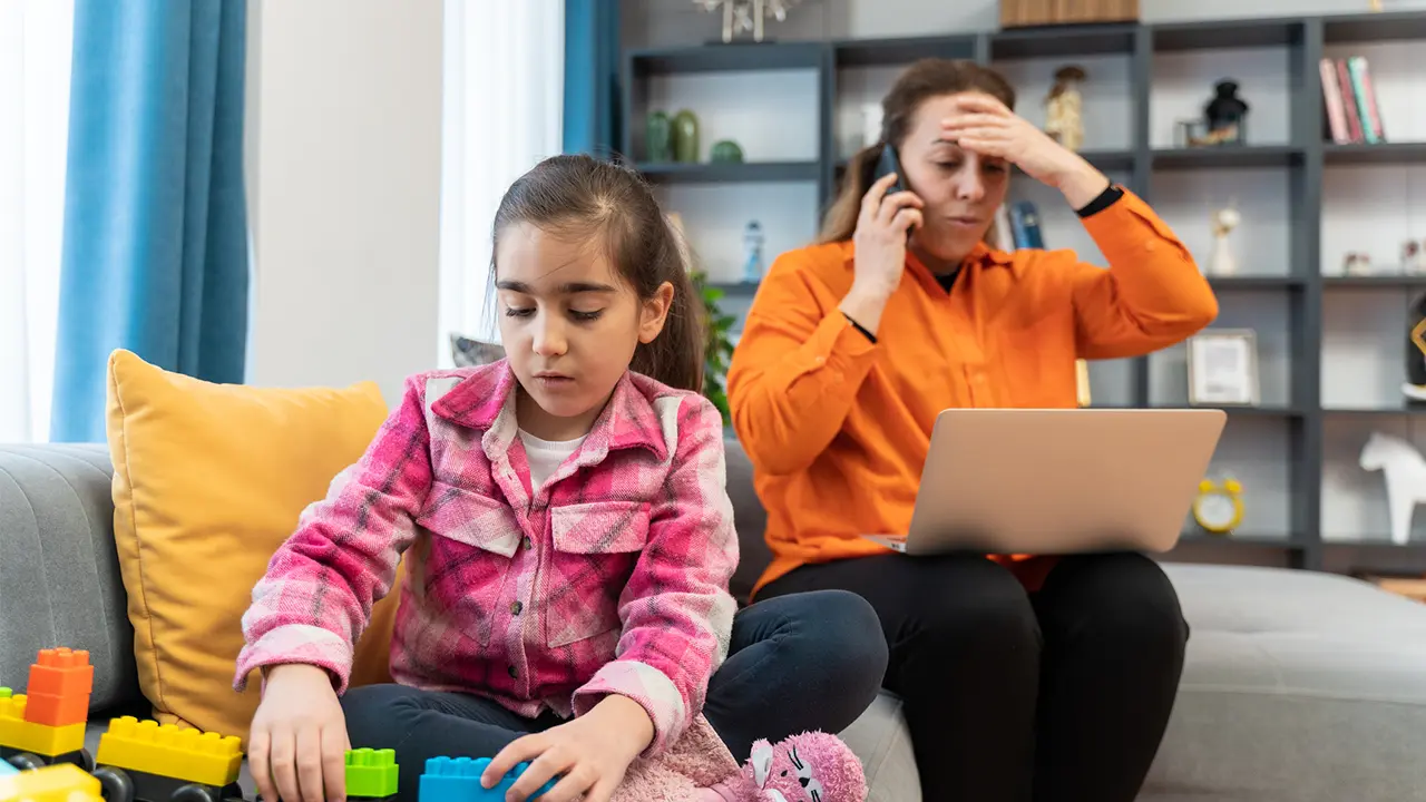 A concerned mother on the phone with a laptop open while her young daughter plays nearby, representing a parent dealing with child identity theft.