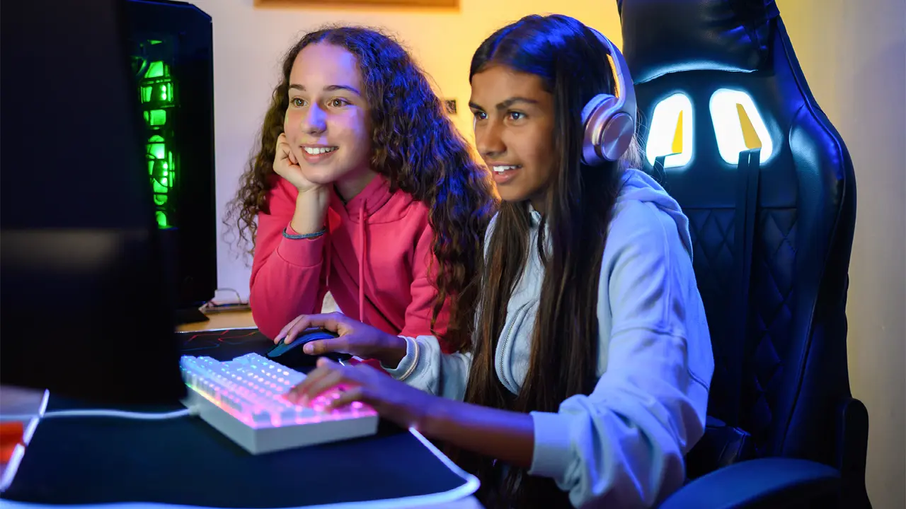 Two girls using a desktop computer with gaming setup, wearing headphones and smiling while gaming online