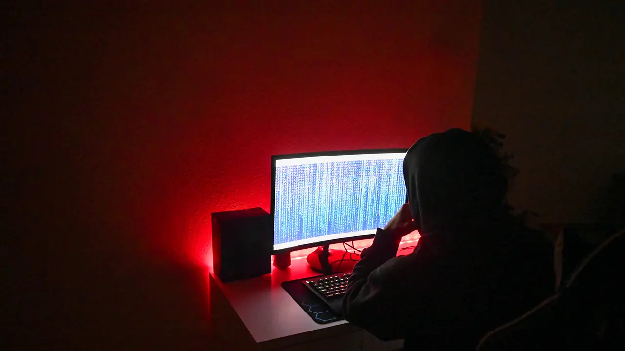 A hooded figure at a computer screen in a dark room with red lighting, representing ghost student loan fraud and identity theft scams targeting federal financial aid.