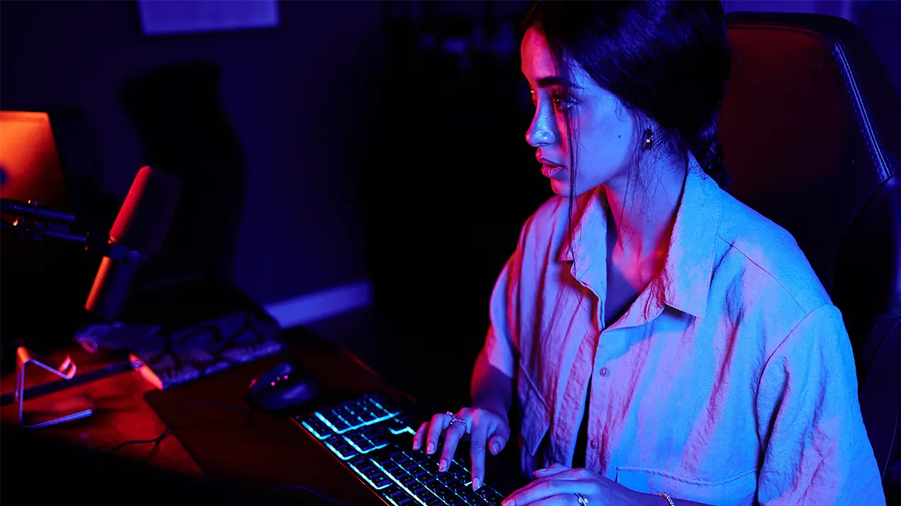 A woman typing at a gaming keyboard in a dark room with blue and red lighting, representing the risk of gaming account breaches and compromised personal data.