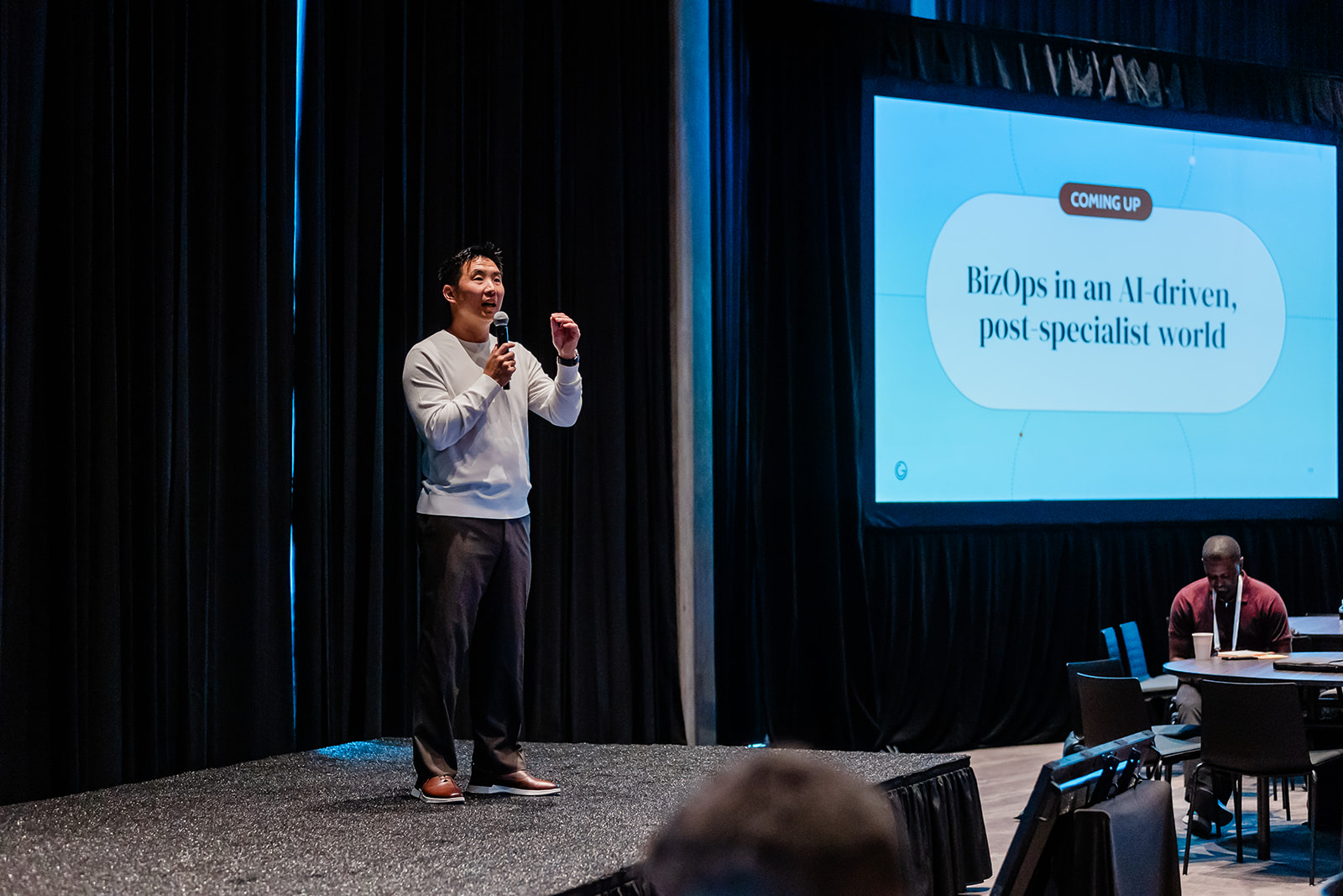 Speaker on stage holding microphone with screen showing BizOps in an AI-driven, post-specialist world.