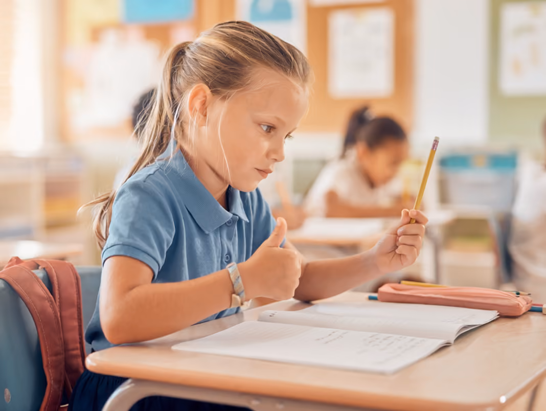 Child sitting at a desk in classroom, holding pencil and giving a thumbs-up while working in notebook.