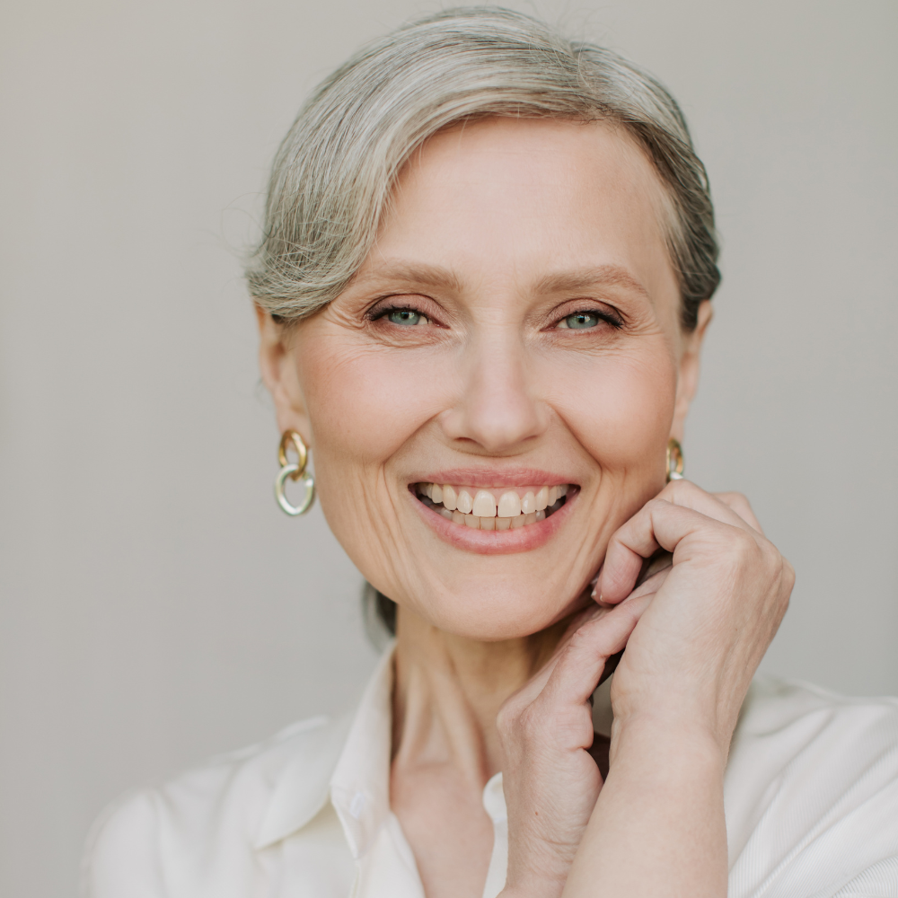 Smiling mature woman with grey hair and blue eyes wearing gold hoop earrings and a white shirt, touching her face.