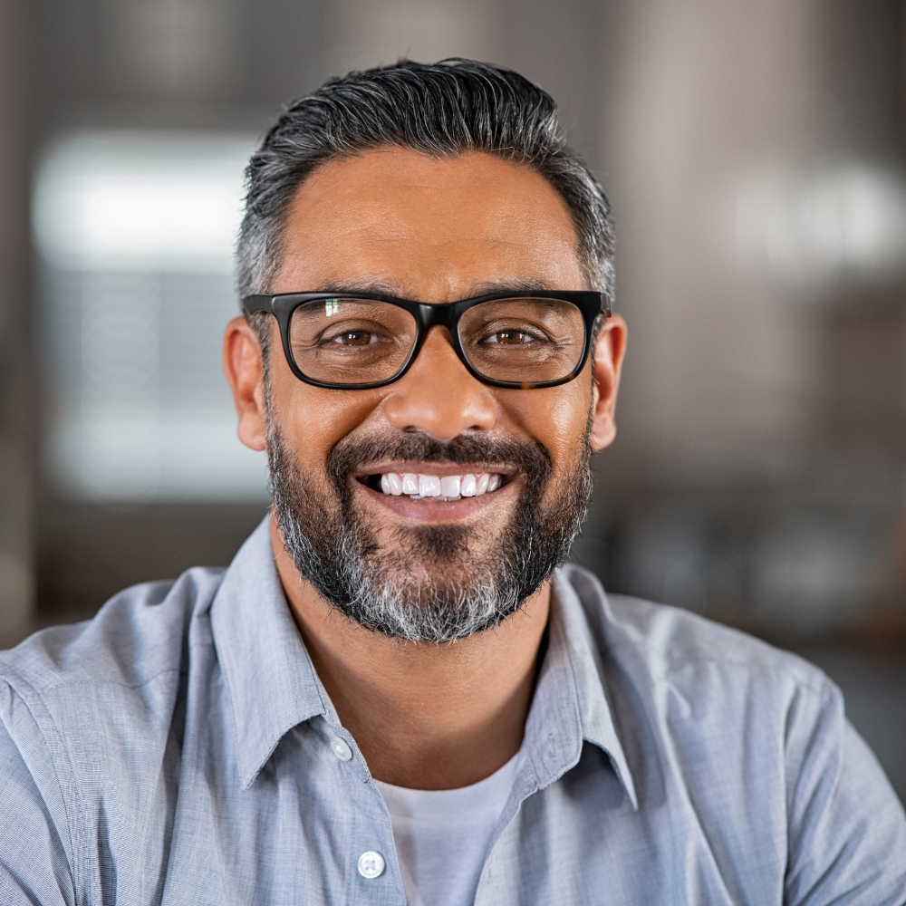 Smiling man with black glasses, gray beard, and short black hair wearing a light blue shirt and white undershirt.